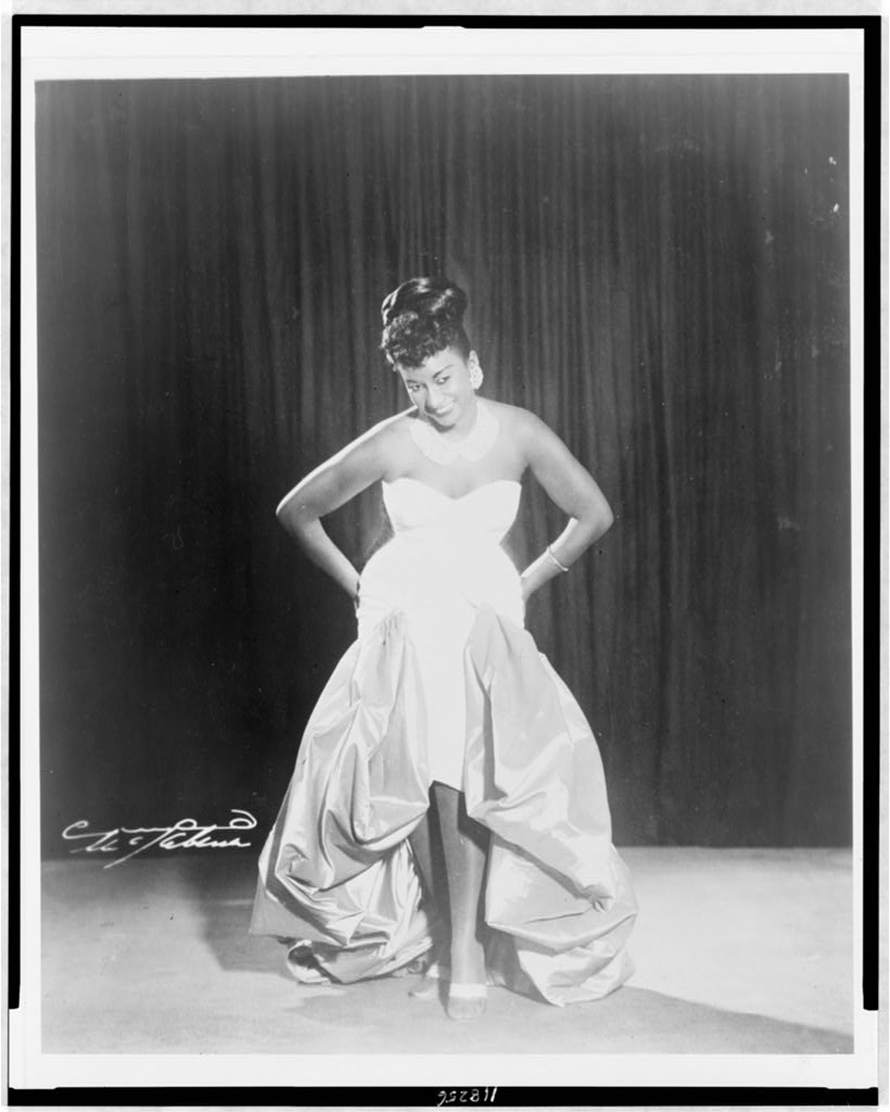 Black and white 1962 photograph of Celia Cruz on stage in a strapless white gown, hands on her hips, smiling down, against a dark curtain, for a post on Celia Cruz and her music in exile